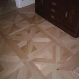 Ułożony parkiet z jasnego drewna w geometryczny wzór, widoczny fragment ciemnego kredensu z szufladami, wzorzysta tapeta w tle.