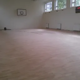 Jasna, odnowiona podłoga parkietowa w sali gimnastycznej z widocznym parkietem ułożonym w cegiełkę, czarną tablicą wyników i koszem do koszykówki na ścianie.