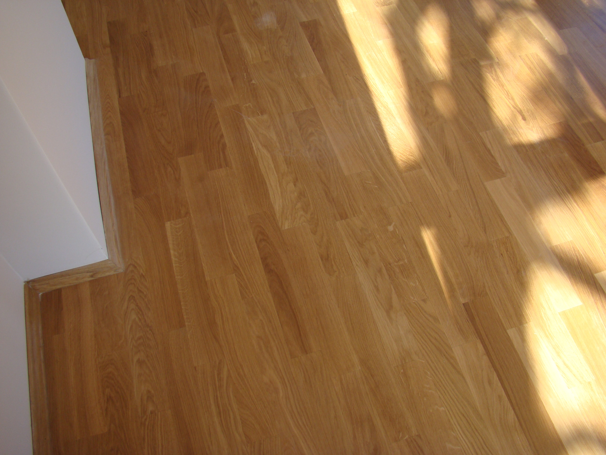 Jasny parkiet dębowy ułożony przy białej ścianie, oświetlony promieniami słonecznymi tworzącymi wyraźne cienie.