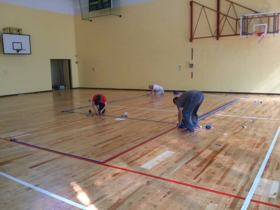 Trzech mężczyzn w hali sportowej, przygotowujących drewnianą podłogę do malowania linii boiska, widoczne taśmy malarskie, puszki i narzędzia.