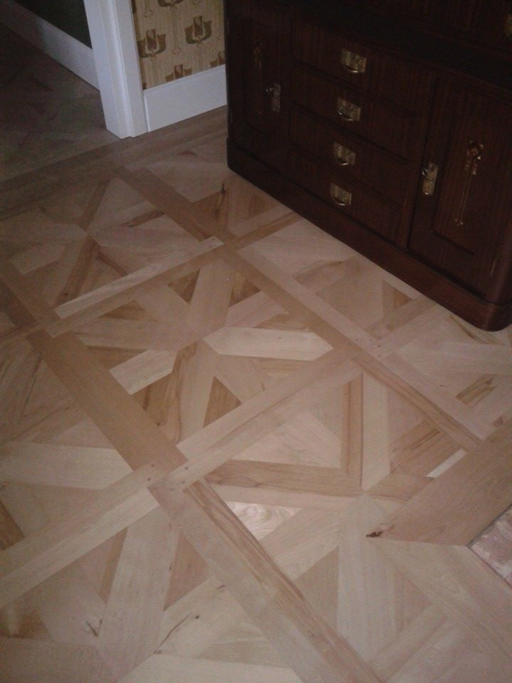 Ułożony parkiet z jasnego drewna w geometryczny wzór, widoczny fragment ciemnego kredensu z szufladami, wzorzysta tapeta w tle.