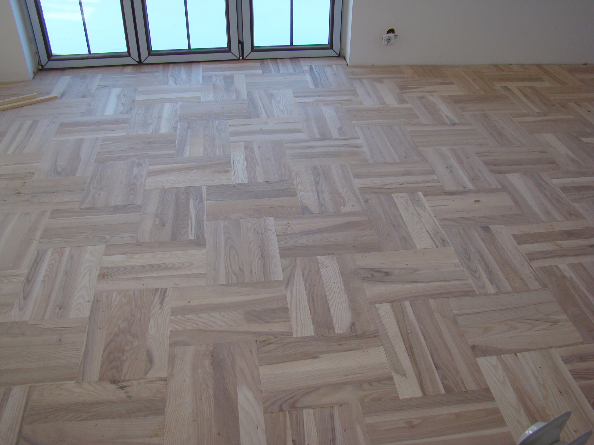 Ułożona parkietowa podłoga w jasnym odcieniu drewna, wzór w kwadraty, widok z góry na fragment pomieszczenia z oknem dachowym i fragmentem ściany z gniazdkiem elektrycznym.