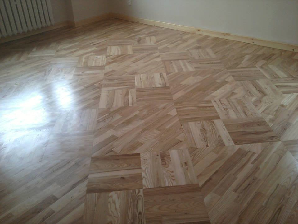 Ułożony parkiet z jasnego drewna w geometryczny wzór kwadratów, widoczne odbicie światła, listwa przypodłogowa i fragment grzejnika.