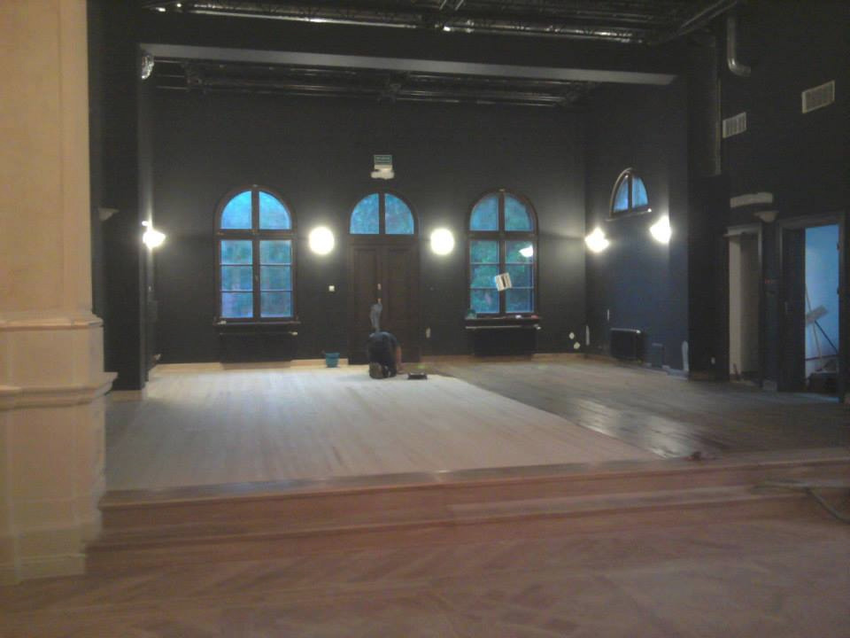 Duża sala z czarnymi ścianami, gdzie część drewnianej podłogi jest już wyszlifowana, a reszta czeka na odnowienie. Widoczna osoba pracująca przy podłodze oraz okna łukowe z niebieskawym światłem...