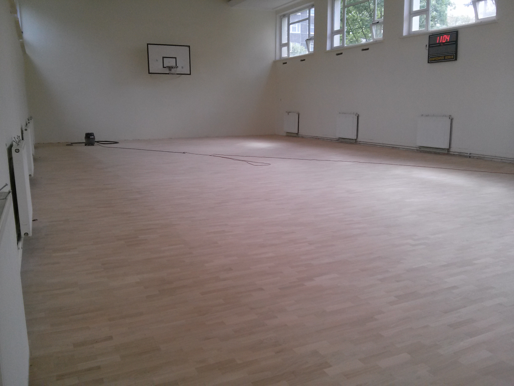 Jasna, odnowiona podłoga parkietowa w sali gimnastycznej z widocznym parkietem ułożonym w cegiełkę, czarną tablicą wyników i koszem do koszykówki na ścianie.