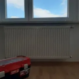 Biały grzejnik panelowy pod oknem z widokiem na błękitne niebo i chmury, obok skrzynka z narzędziami na drewnianej podłodze.