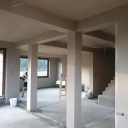 Wnętrze domu w trakcie tynkowania maszynowego, widoczne słupy konstrukcyjne i ściany pokryte świeżym tynkiem, przewody elektryczne na podłodze, drabina w tle i osoba w oknie.