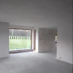 Wnętrze domu w stanie deweloperskim po wylewce posadzki, z widokiem na ogród przez duże okno. Ściany przygotowane pod malowanie, widoczne puszki elektryczne.