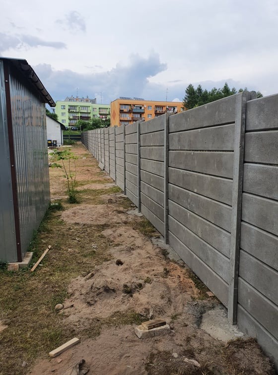 Długi, szary mur z prefabrykatów w Brody, z widocznymi poziomymi panelami i słupkami, oddzielający posesję od sąsiedniej działki z budynkami mieszkalnymi w tle.