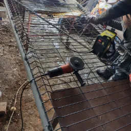 Spawanie metalowej konstrukcji ogrodzeniowej z użyciem żółtej spawarki inwertorowej, widoczna szlifierka kątowa i iskry podczas spawania.