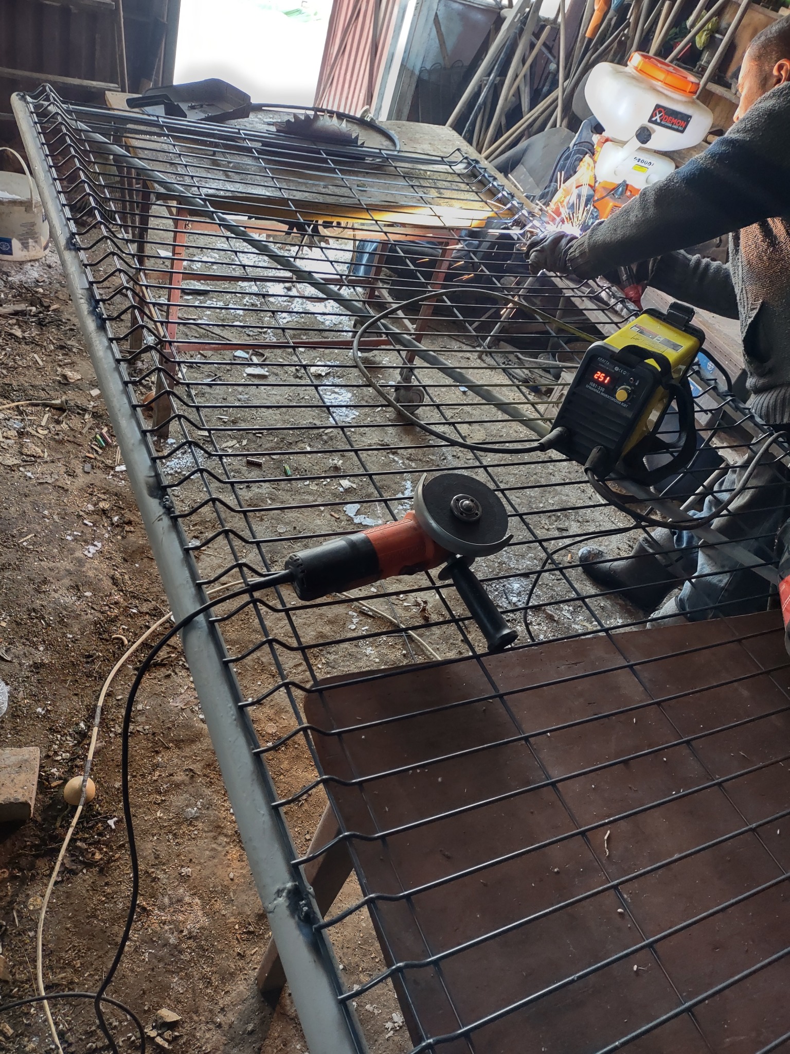 Spawanie metalowej konstrukcji ogrodzeniowej z użyciem żółtej spawarki inwertorowej, widoczna szlifierka kątowa i iskry podczas spawania.