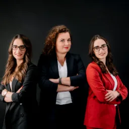 Portret trzech uśmiechniętych kobiet w eleganckich, kolorowych marynarkach na ciemnym tle. Kobiety mają założone okulary i skrzyżowane ręce.