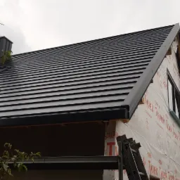 Fragment domu w trakcie prac budowlanych z nowym, czarnym dachem z dachówki ceramicznej, widoczny komin i okno, elewacja zabezpieczona folią budowlaną, drabina oparta o ścianę.