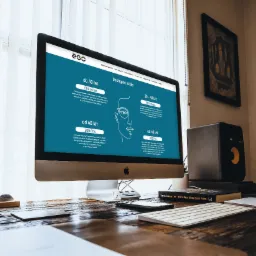 Monitor iMac wyświetlający stronę internetową o szkłach okularowych na drewnianym biurku z akcesoriami, w tle okno z żaluzjami i obraz na ścianie.
