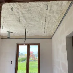 Poddasze po natrysku pianki izolacyjnej, widoczne metalowe uchwyty mocujące, surowy stan ścian, okno z widokiem na ogród.