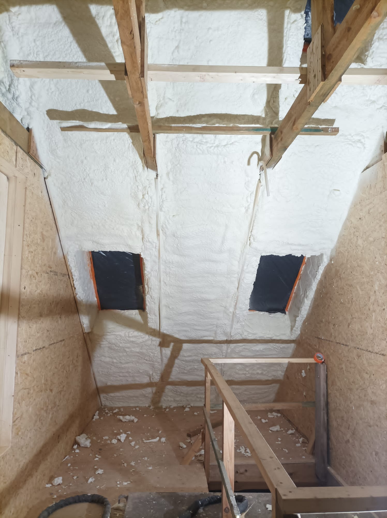 Poddasze w Zduńskiej Woli po aplikacji pianki poliuretanowej, widoczne drewniane krokwie i dwa okna dachowe zabezpieczone folią, na podłodze resztki pianki.