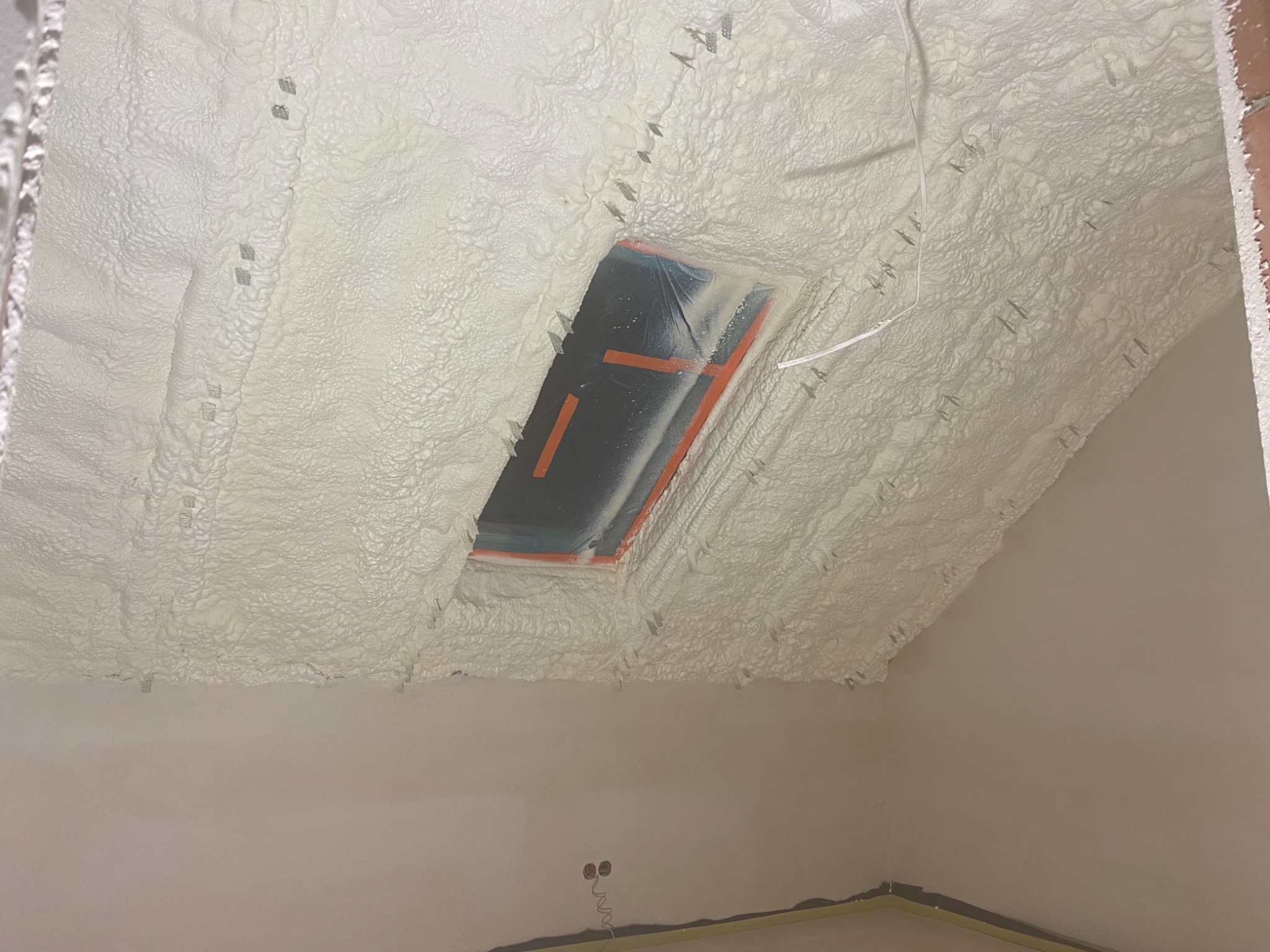 Poddasze z oknem dachowym po natrysku izolacji pianką PUR, widoczne metalowe elementy mocujące i folia zabezpieczająca okno.