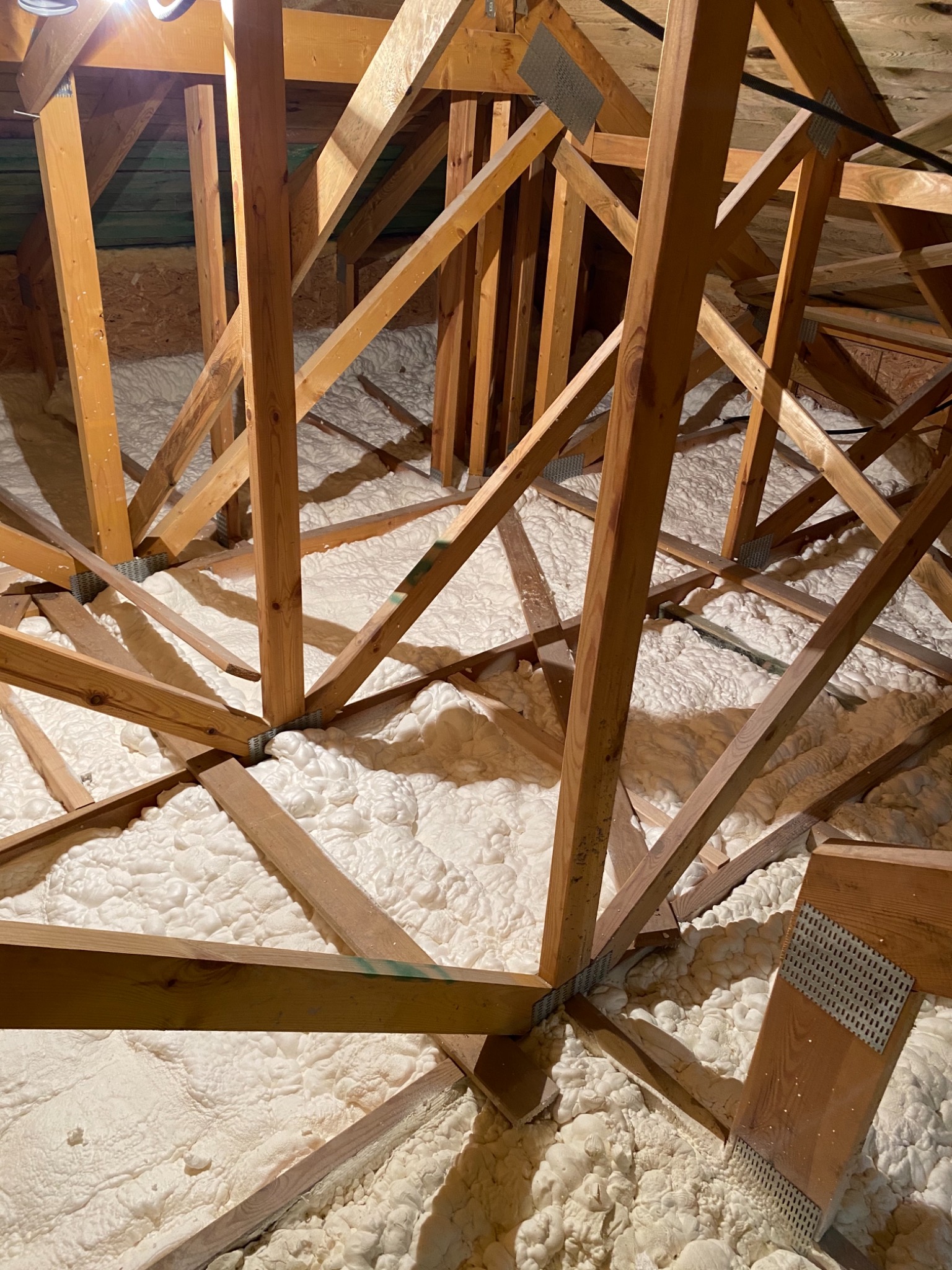 Poddasze z drewnianą konstrukcją krokwiową wypełnione białą pianą izolacyjną, widoczne metalowe łączniki i fragmenty zielonej impregnacji drewna.
