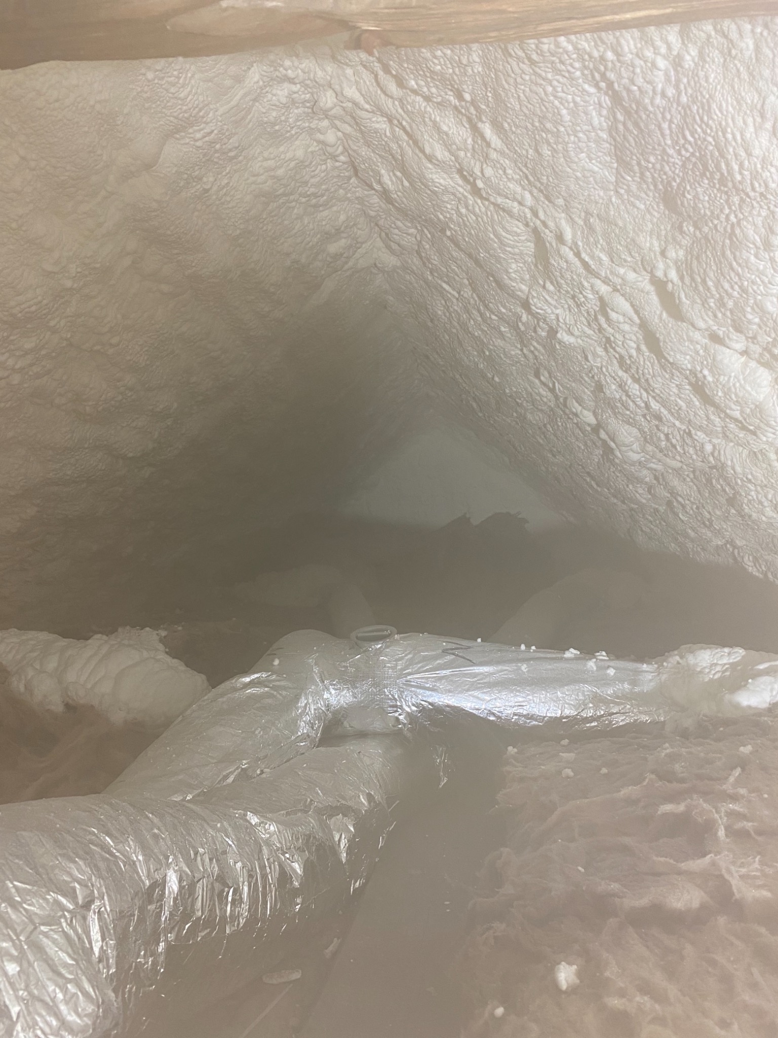 Poddasze z widoczną warstwą białej pianki izolacyjnej, pokrywającej skośne powierzchnie dachu i fragment przewodu wentylacyjnego w srebrnej folii.