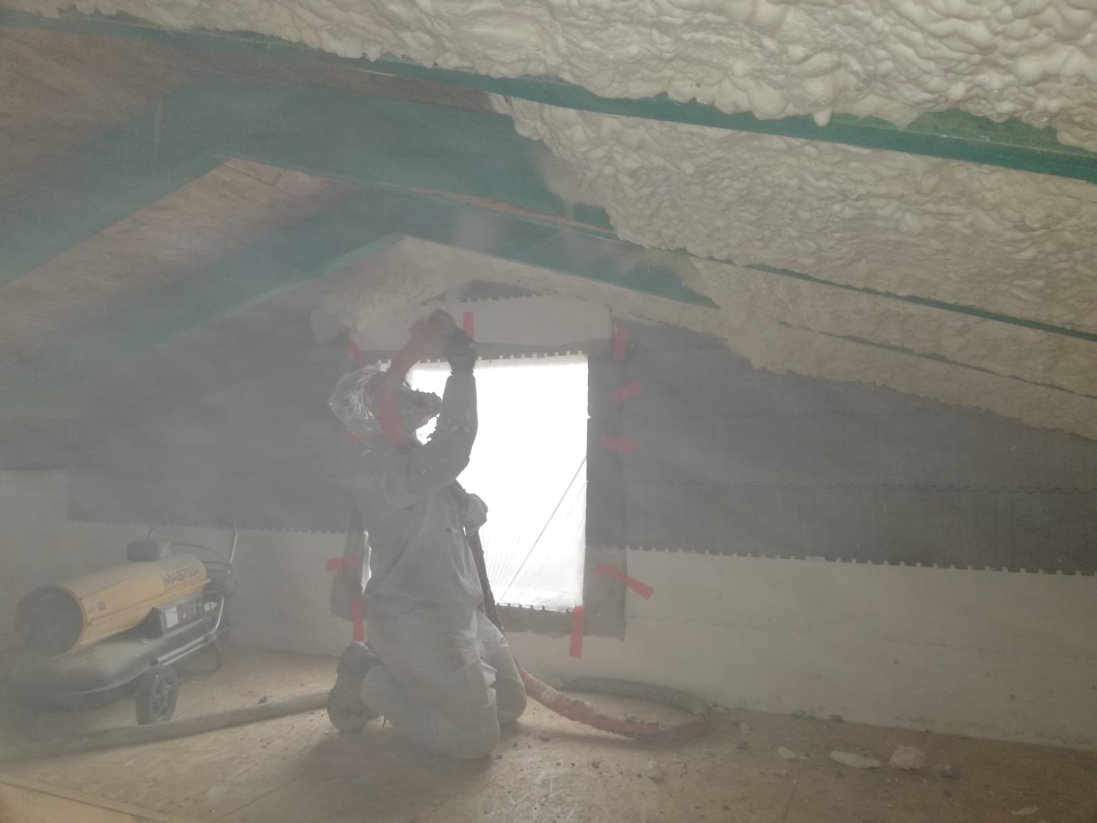 Pracownik w kombinezonie ochronnym aplikuje pianę izolacyjną na poddaszu, widoczny agregat natryskowy Master, okno zabezpieczone folią, częściowo zaizolowany dach z widocznymi elementami...