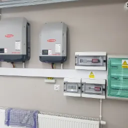Dwa szare inwertery Fronius Symo zamontowane na ścianie nad grzejnikiem, obok rozdzielnice elektryczne w białych obudowach. Widoczny fragment instalacji elektrycznej z przewodami i gniazdkiem.