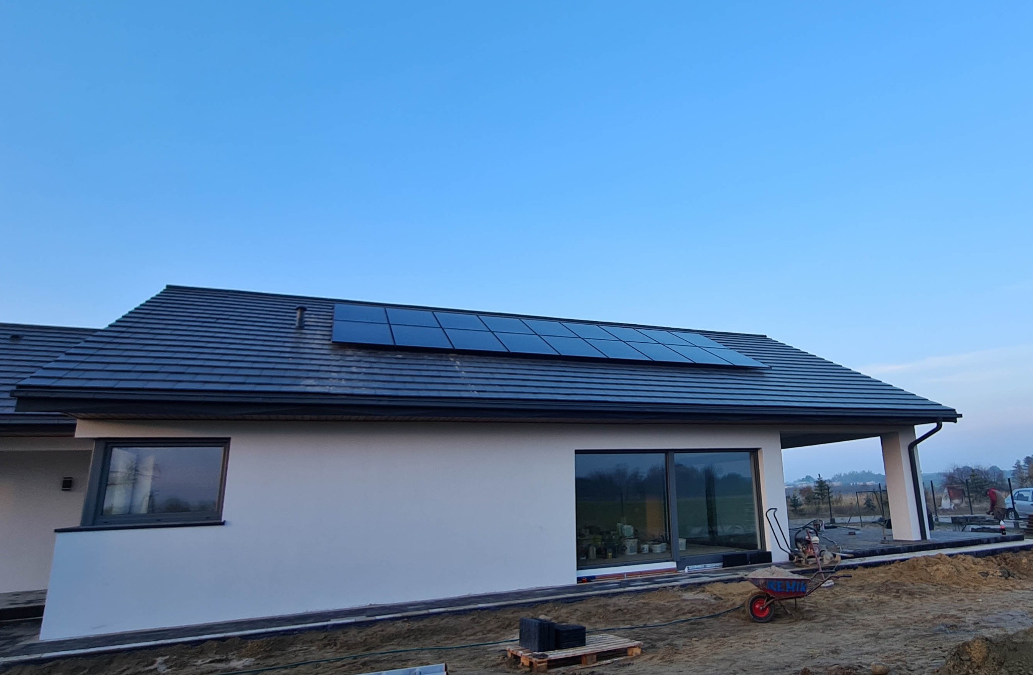 Nowoczesny dom jednorodzinny z panelami słonecznymi na grafitowym dachu, widoczne okna z ciemnymi ramami i teren w trakcie prac wykończeniowych.