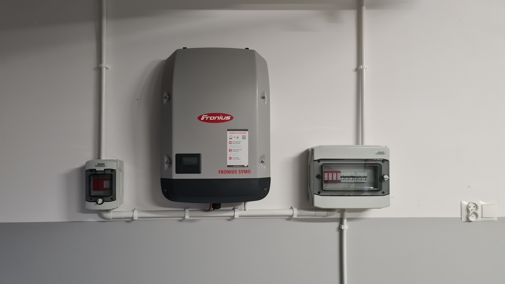 Szary inwerter Fronius Symo zamontowany na białej ścianie, obok skrzynki zabezpieczeń i gniazdka elektrycznego, kable w białych listwach.