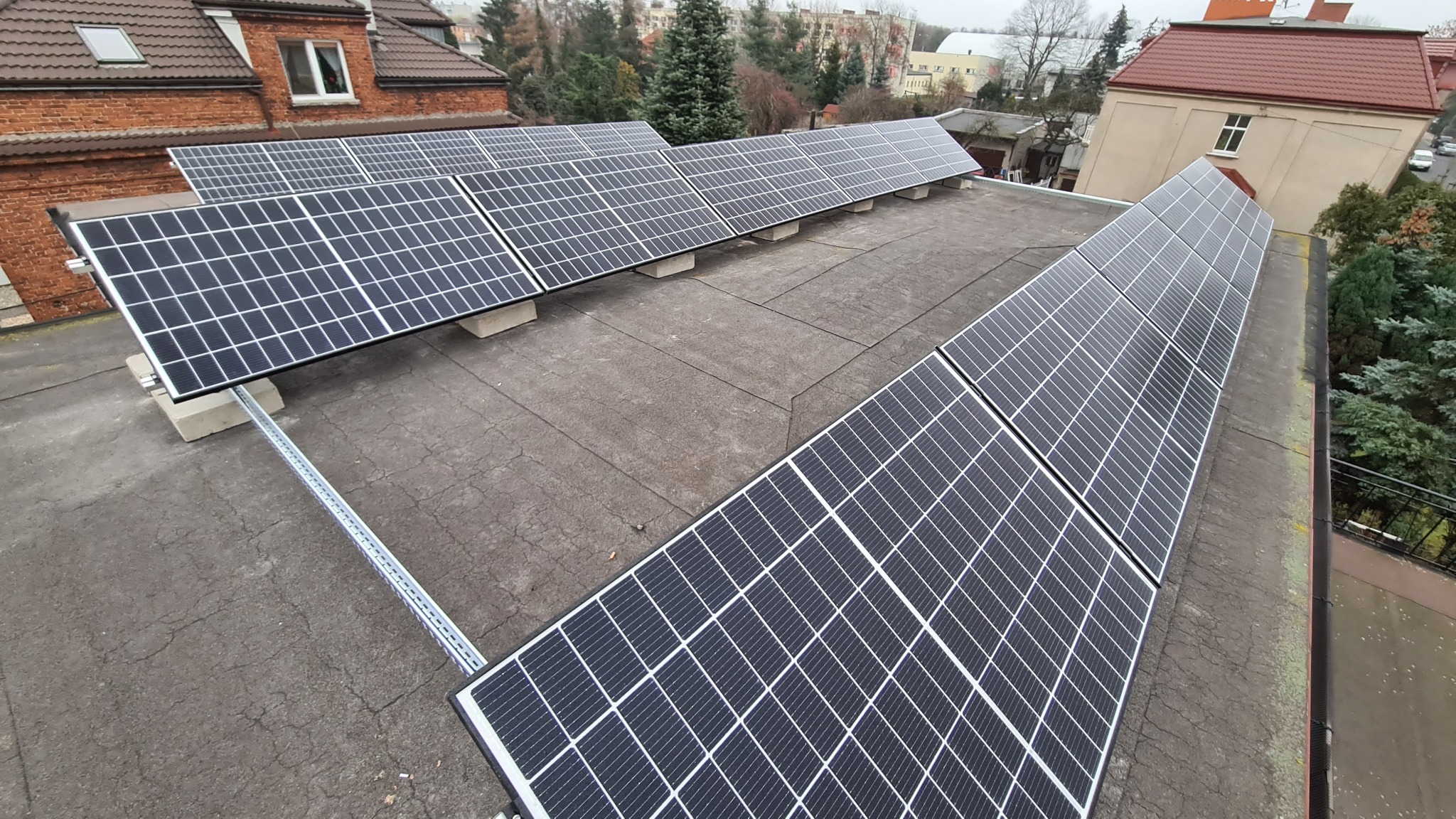 Instalacja paneli słonecznych na płaskim dachu budynku z widocznymi elementami montażowymi i betonowymi podstawami, otoczenie miejskie z budynkami i drzewami w tle.