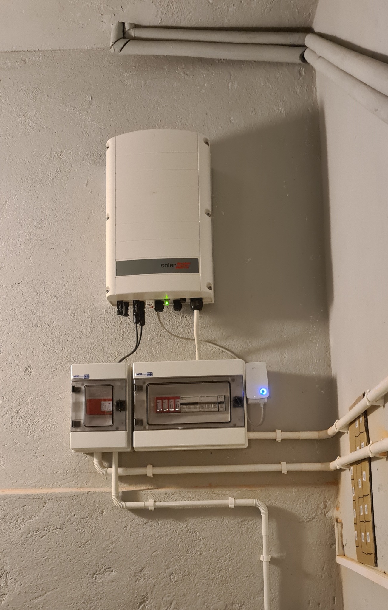 Inwerter solarny z zieloną diodą LED, podłączony do dwóch skrzynek bezpiecznikowych i wzmacniacza sygnału Wi-Fi, zamontowane na szarej, surowej ścianie. Widoczne rury instalacyjne.