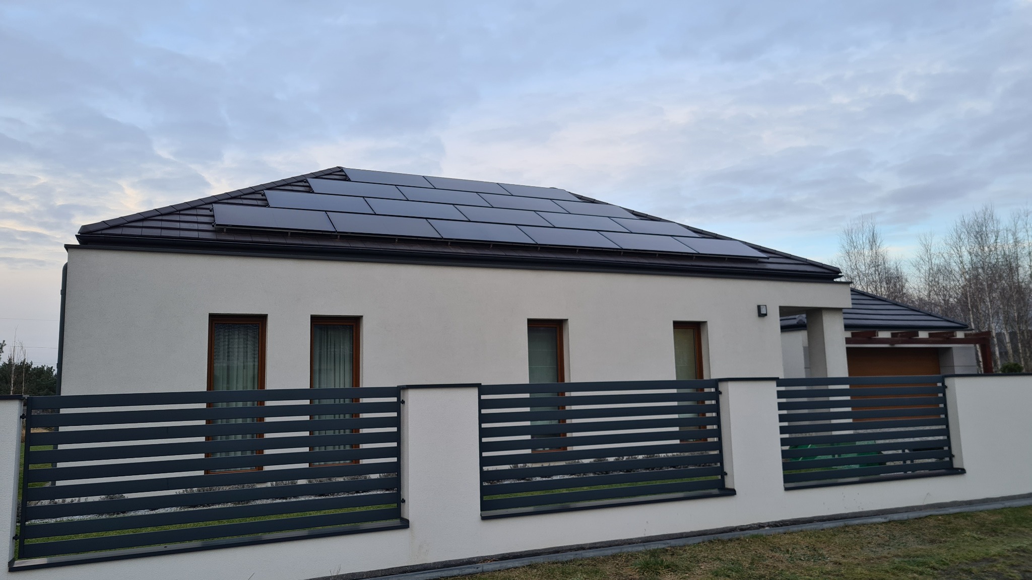 Nowoczesny dom jednorodzinny z panelami słonecznymi na dachu, widoczne grafitowe ogrodzenie poziome i trzy okna z brązowymi ramami.