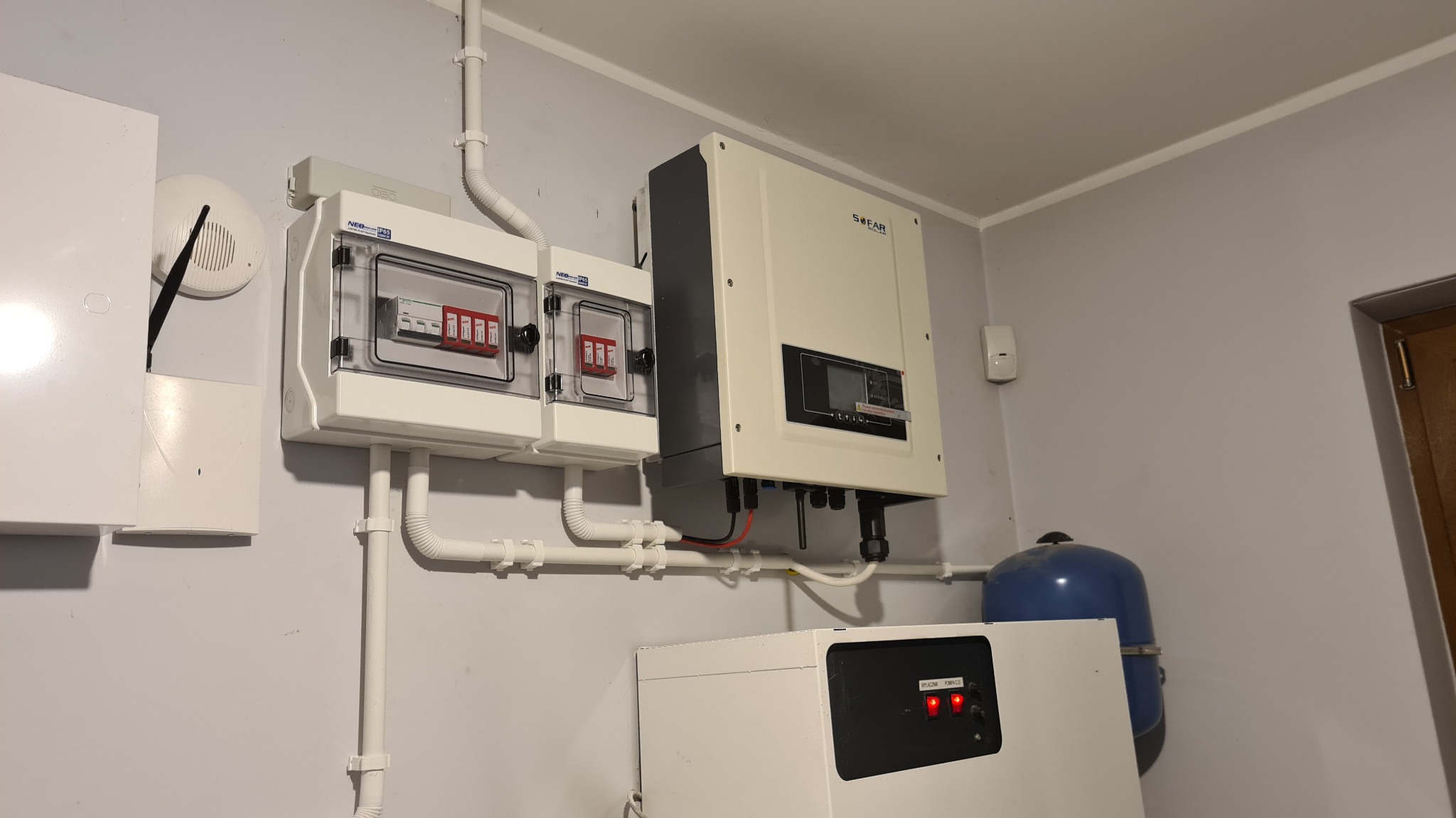 Biała skrzynka elektryczna z bezpiecznikami, inwerter SOFAR i zbiornik ciśnieniowy zamontowane na szarej ścianie, widoczne białe rury instalacyjne i czujnik dymu.