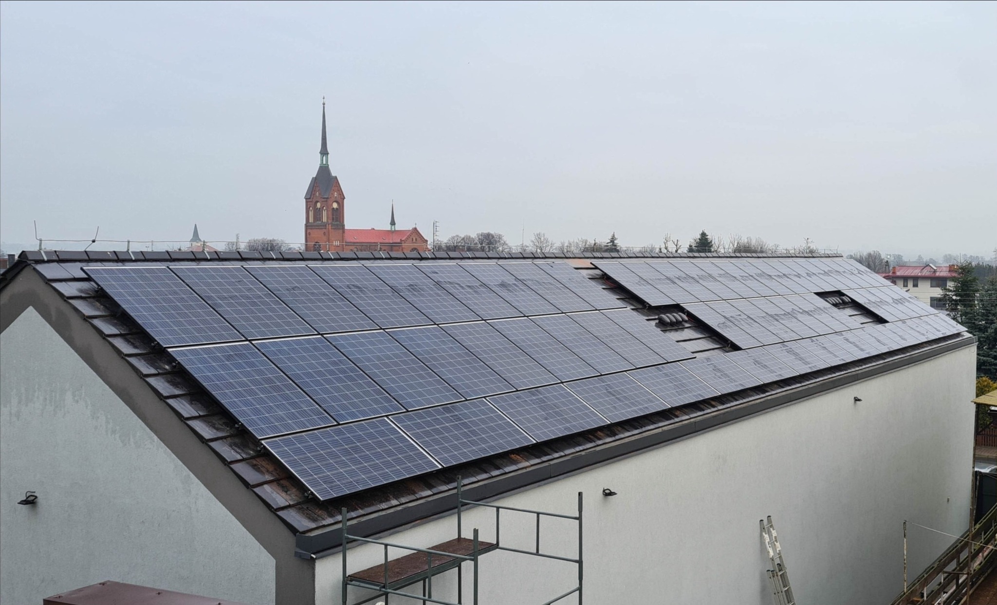 Panele fotowoltaiczne na dachu domu jednorodzinnego, widoczne rusztowanie i drabina, w tle kościół w oddali, pochmurny dzień.