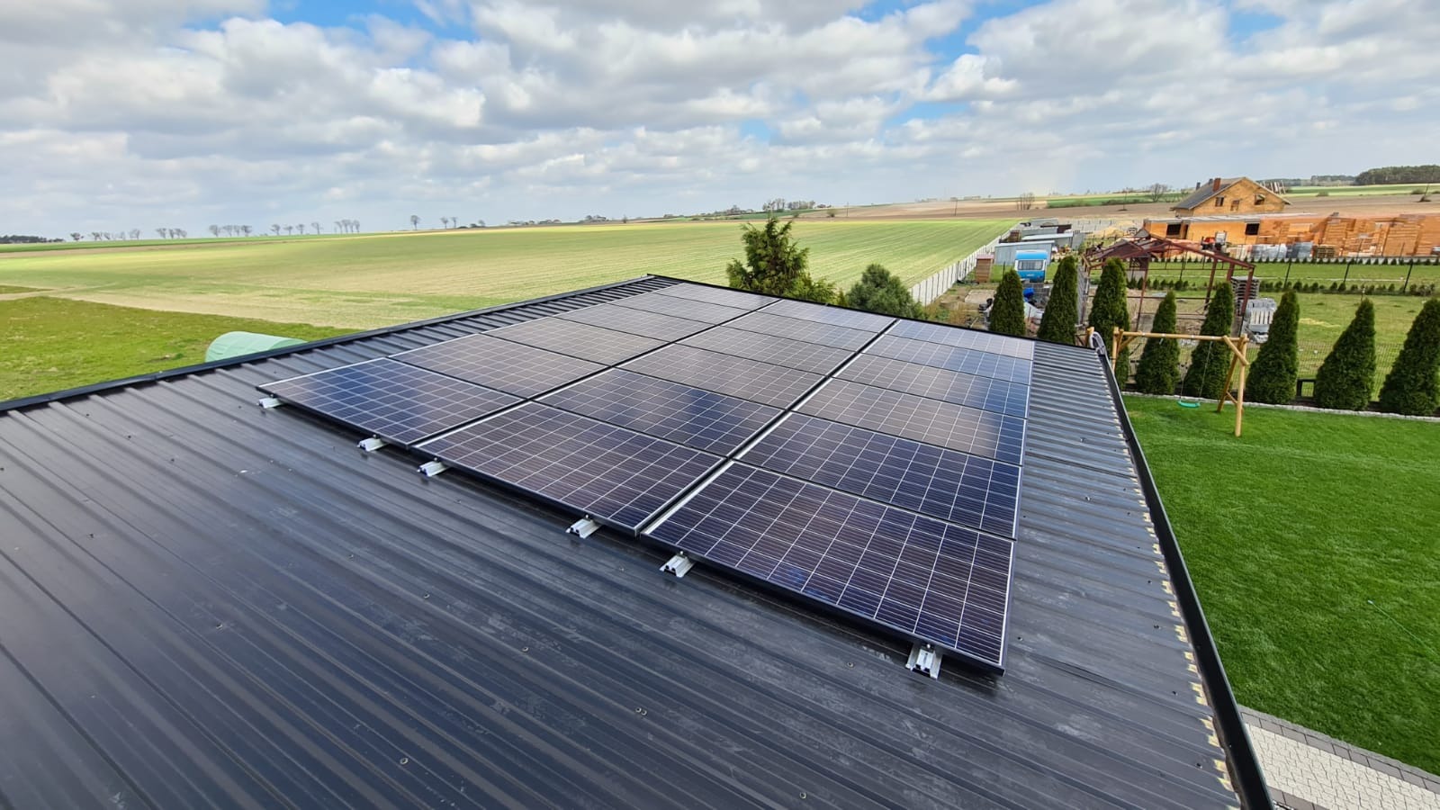 Panele słoneczne na dachu z blachy trapezowej, widok na zielone pola i budowę w tle, błękitne niebo z chmurami.