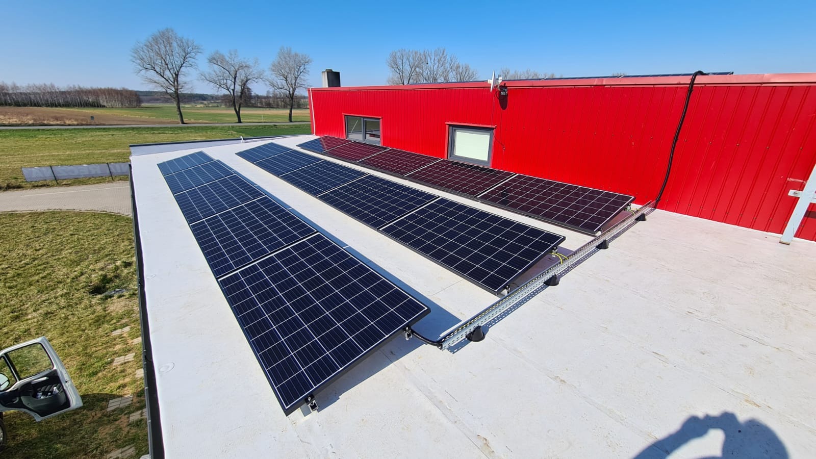 Panele słoneczne zamontowane na białym dachu budynku z czerwoną ścianą. Widoczne elementy montażowe, kable i krajobraz w tle.