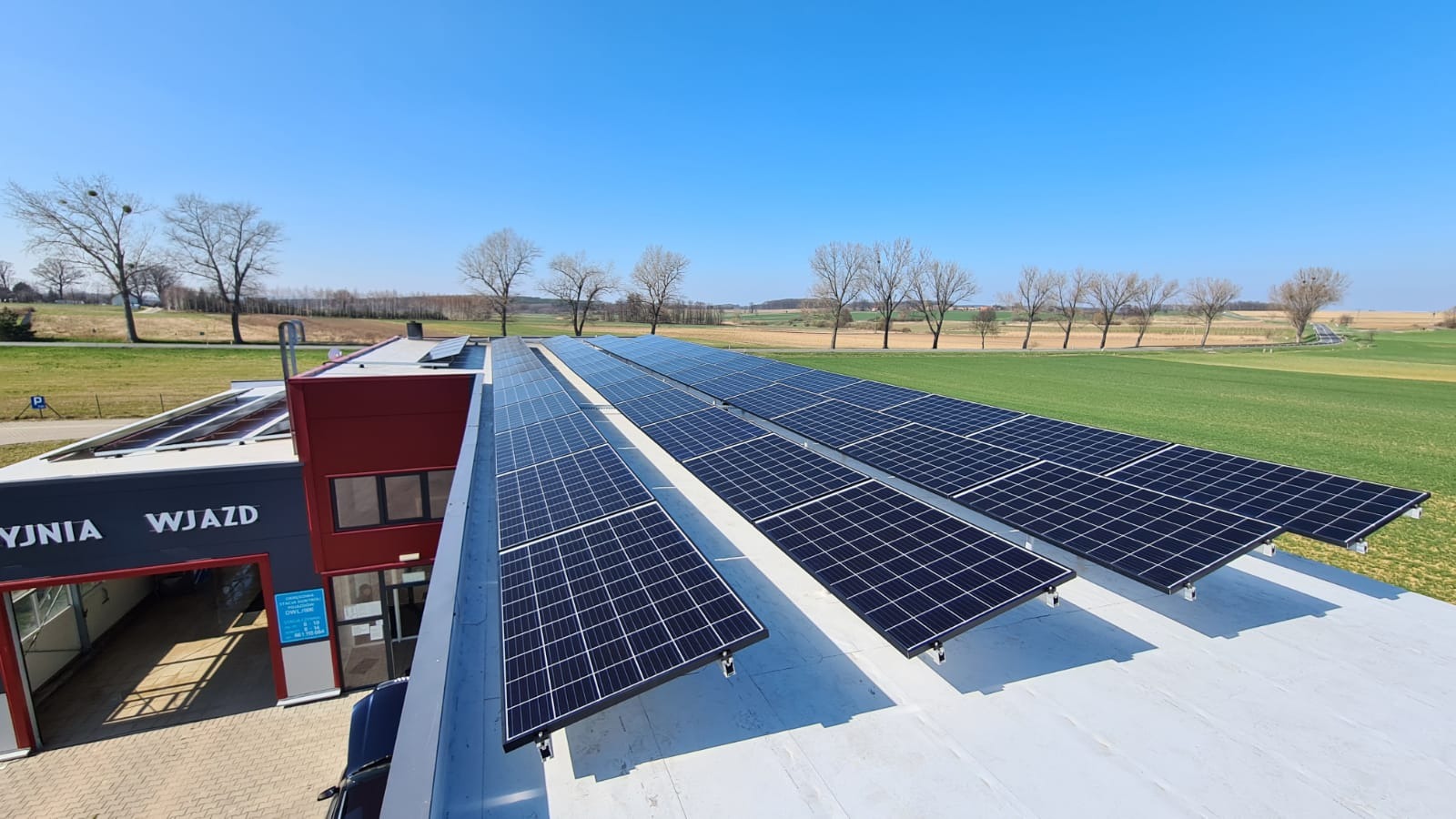 Rząd paneli słonecznych na dachu budynku myjni samochodowej z widokiem na pola i drzewa w słoneczny dzień.