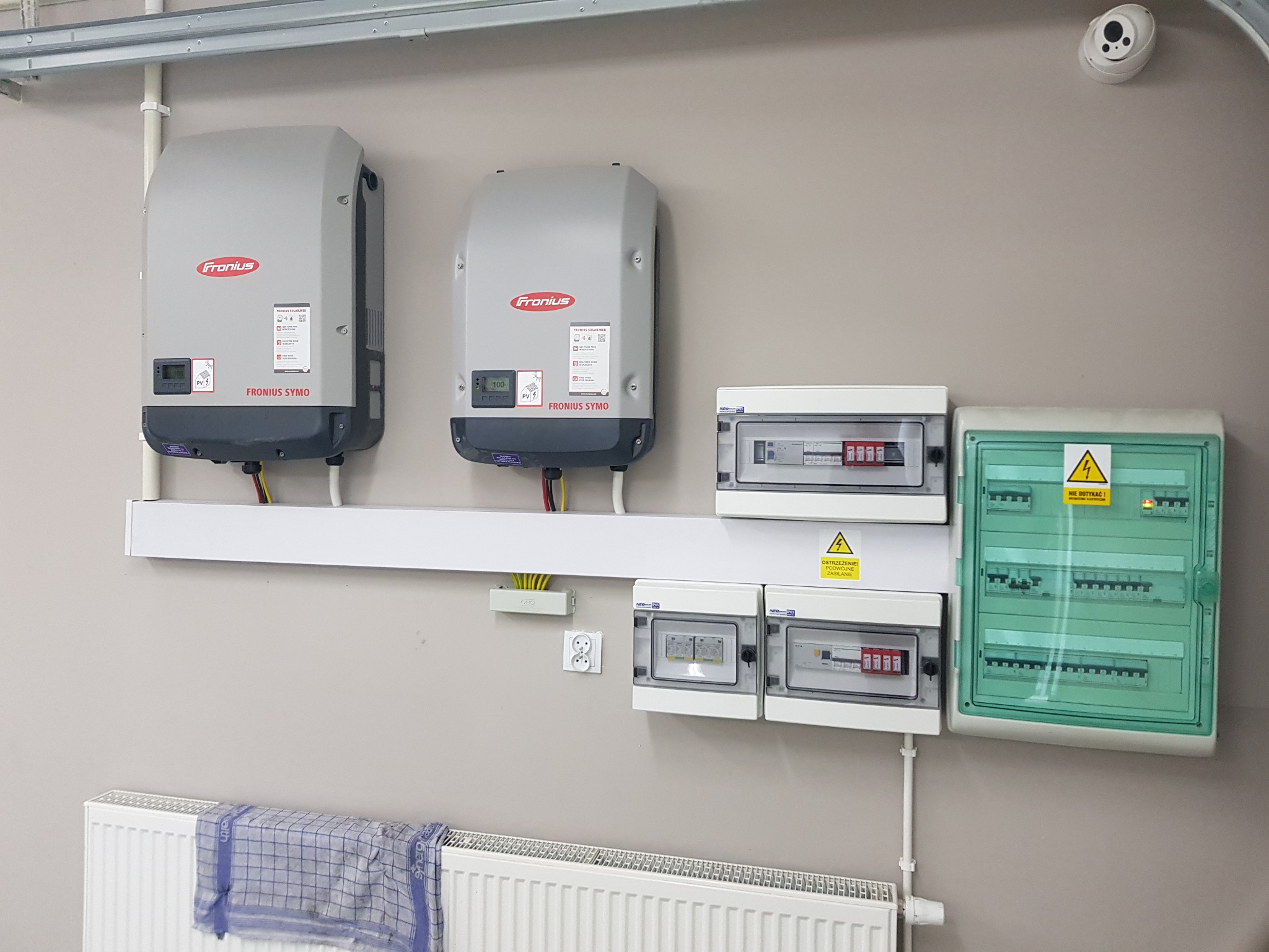 Dwa szare inwertery Fronius Symo zamontowane na ścianie nad grzejnikiem, obok rozdzielnice elektryczne w białych obudowach. Widoczny fragment instalacji elektrycznej z przewodami i gniazdkiem.