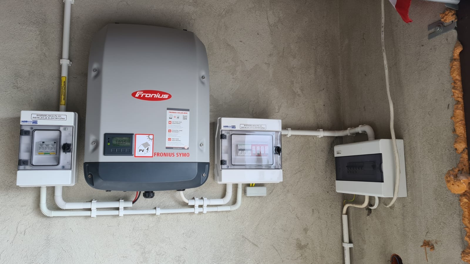 Szary falownik Fronius Symo z wyświetlaczem wskazującym 1835, zamontowany na szarej ścianie obok dwóch białych skrzynek rozdzielczych PV DC i PV AC z widocznymi bezpiecznikami, połączonych białymi...