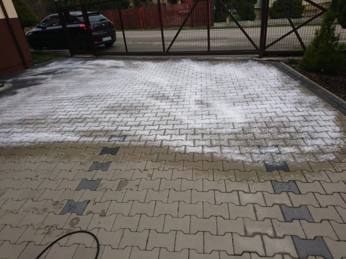 Częściowo oczyszczona z białego nalotu kostka brukowa na podjeździe, widoczna brama w tle i zaparkowany samochód.