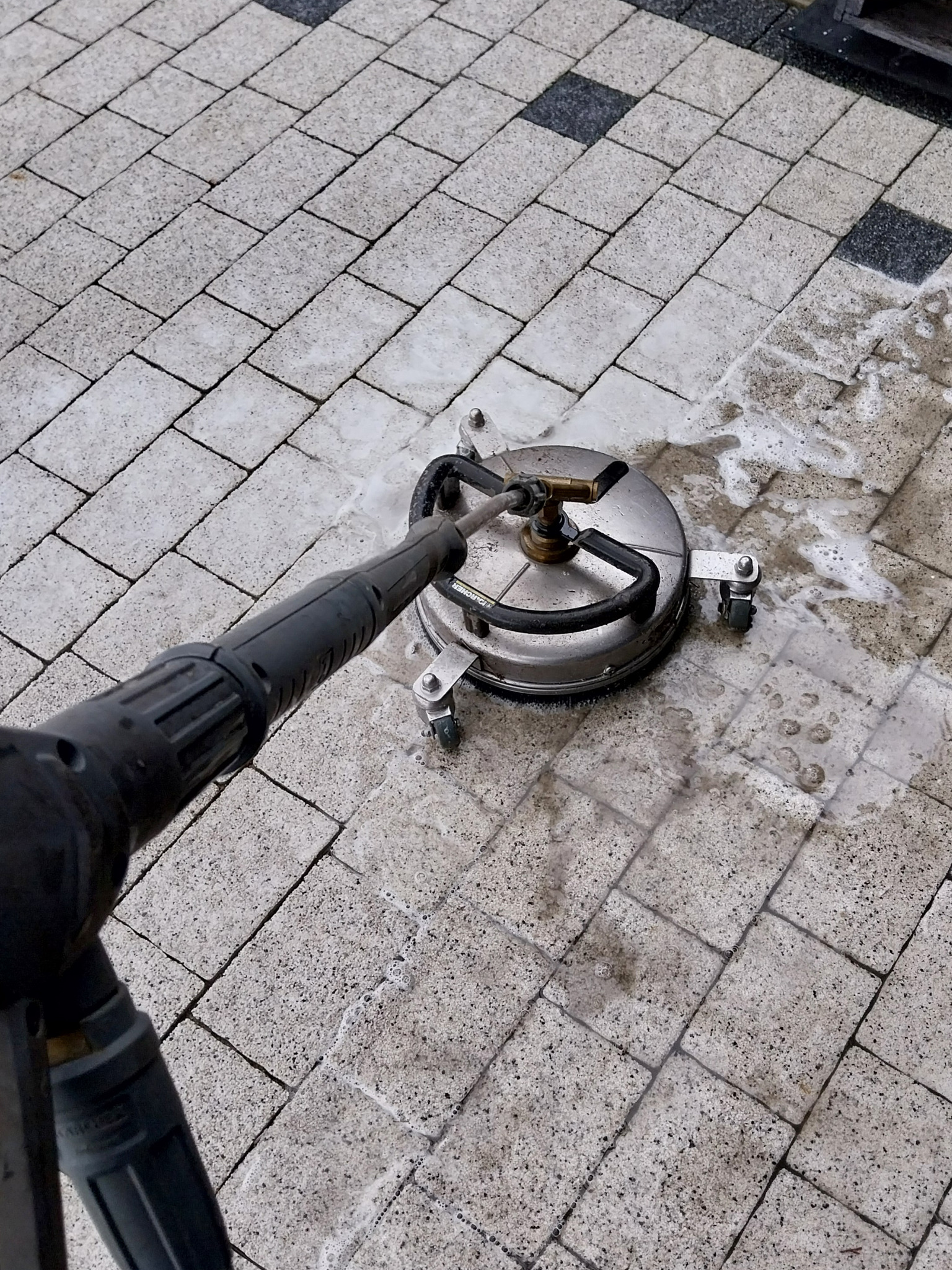 Czyszczenie kostki brukowej szarą myjką ciśnieniową z okrągłą dyszą, widoczne zabrudzenia i piana na czyszczonej powierzchni.