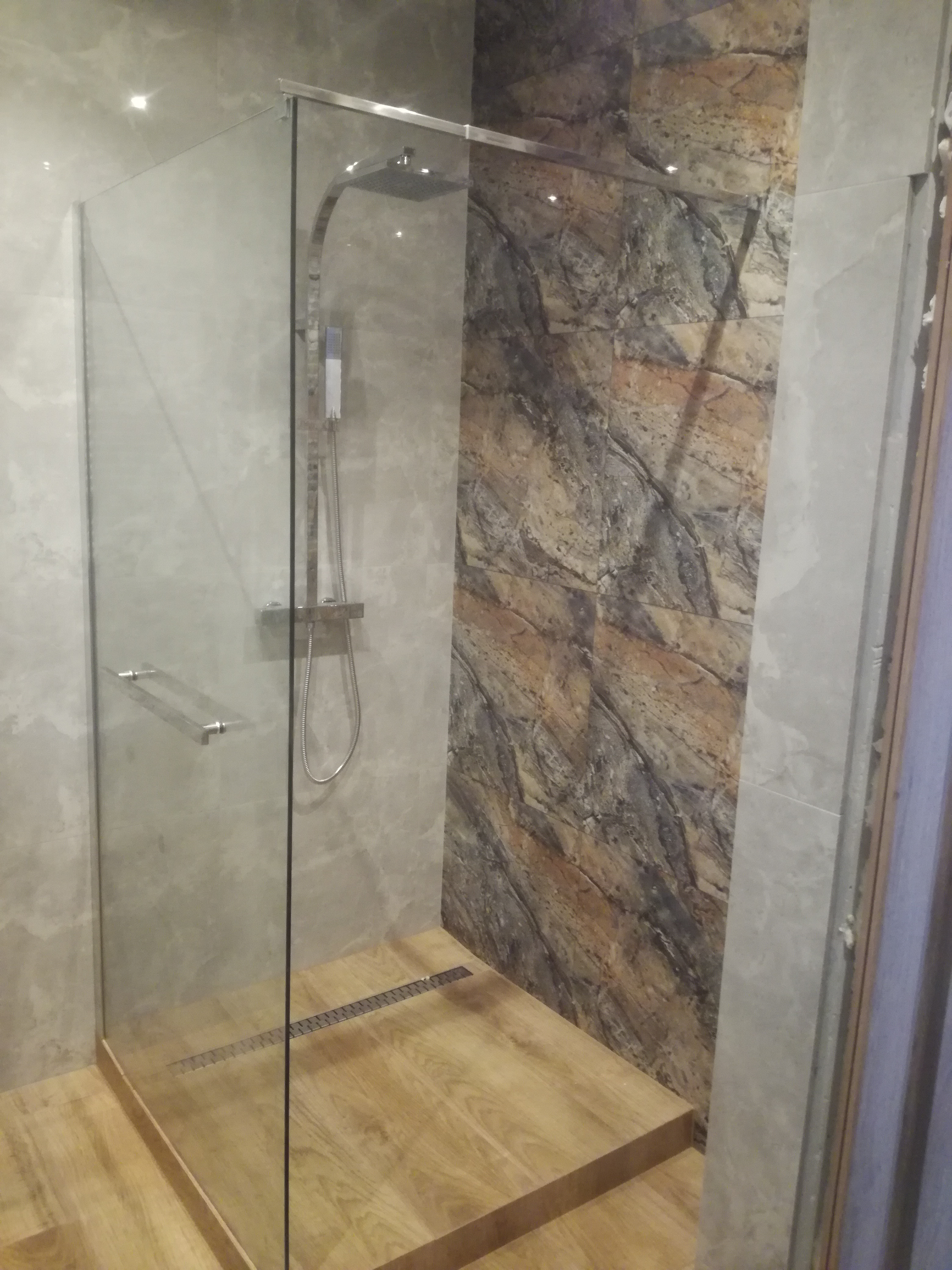 Nowoczesna kabina prysznicowa typu walk-in z drewnianym podestem, odpływem liniowym i panelami ściennymi imitującymi marmur.