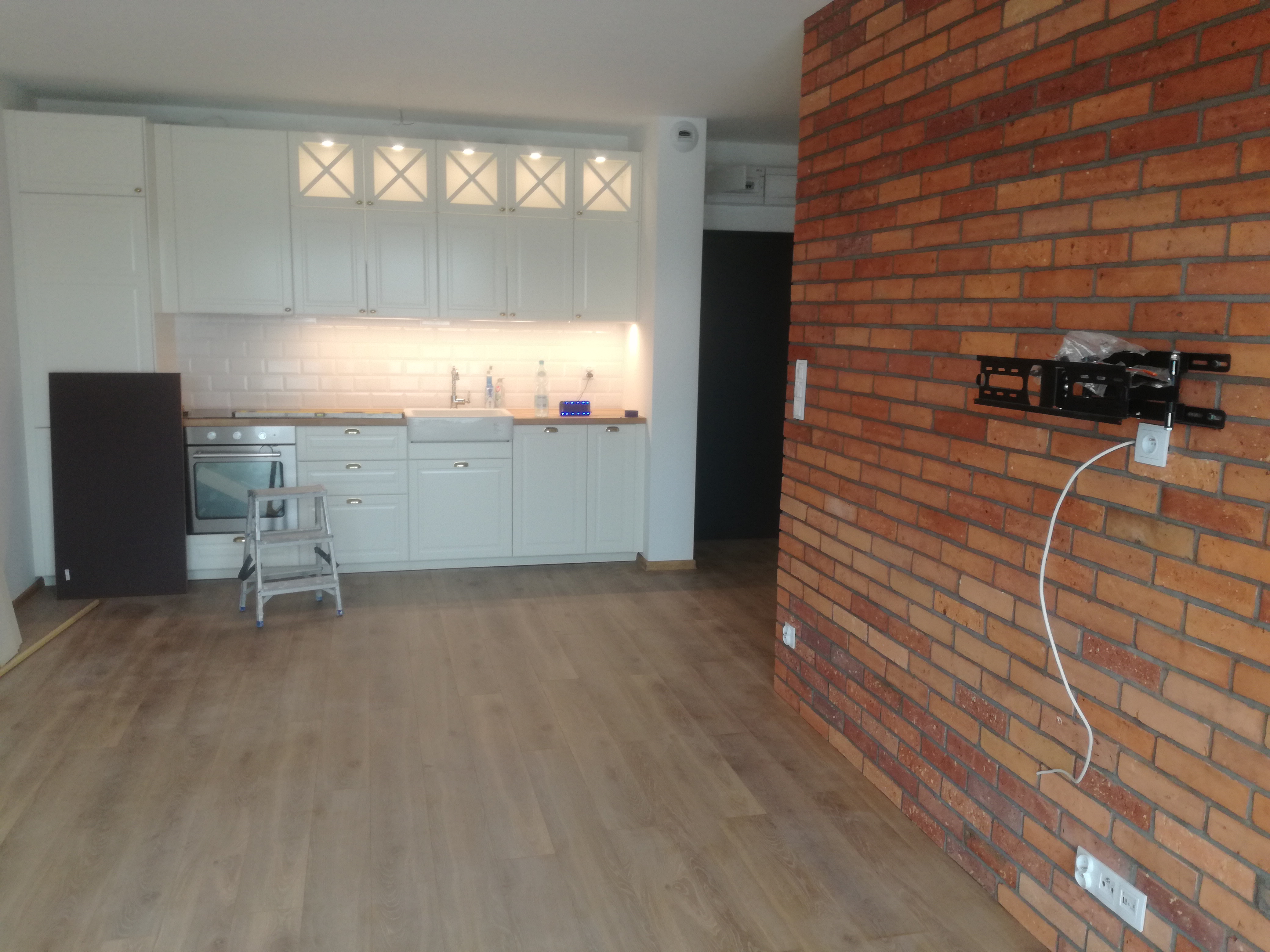 Nowoczesna kuchnia z białymi szafkami, białymi płytkami na ścianie, drewnianym blatem, ceglaną ścianą i zamontowanym uchwytem do telewizora, widoczna drabina malarska.