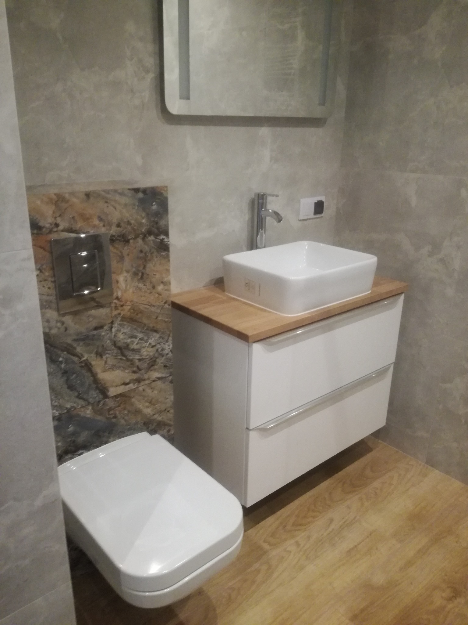 Nowoczesna łazienka w szarościach z drewnianym blatem pod umywalką i marmurową ścianą z podtynkowym WC.