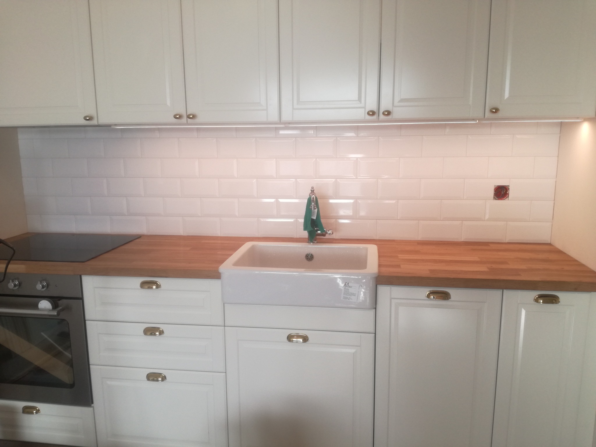 Biała kuchnia z drewnianym blatem, białymi kafelkami typu metro i ceramicznym zlewem farmerskim. Widoczna płyta grzewcza i piekarnik.