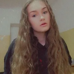 Portret młodej kobiety z długimi, kręconymi włosami w odcieniach blondu i brązu, ubrana w czarną bluzę z delikatnym czerwonym akcentem, na tle jasnych ścian.