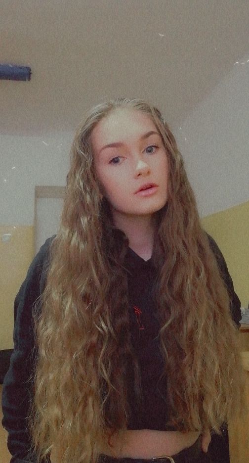 Portret młodej kobiety z długimi, kręconymi włosami w odcieniach blondu i brązu, ubrana w czarną bluzę z delikatnym czerwonym akcentem, na tle jasnych ścian.