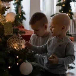 Dwóch chłopców w szarych swetrach dekoruje choinkę bombkami i światełkami w jasnym wnętrzu.