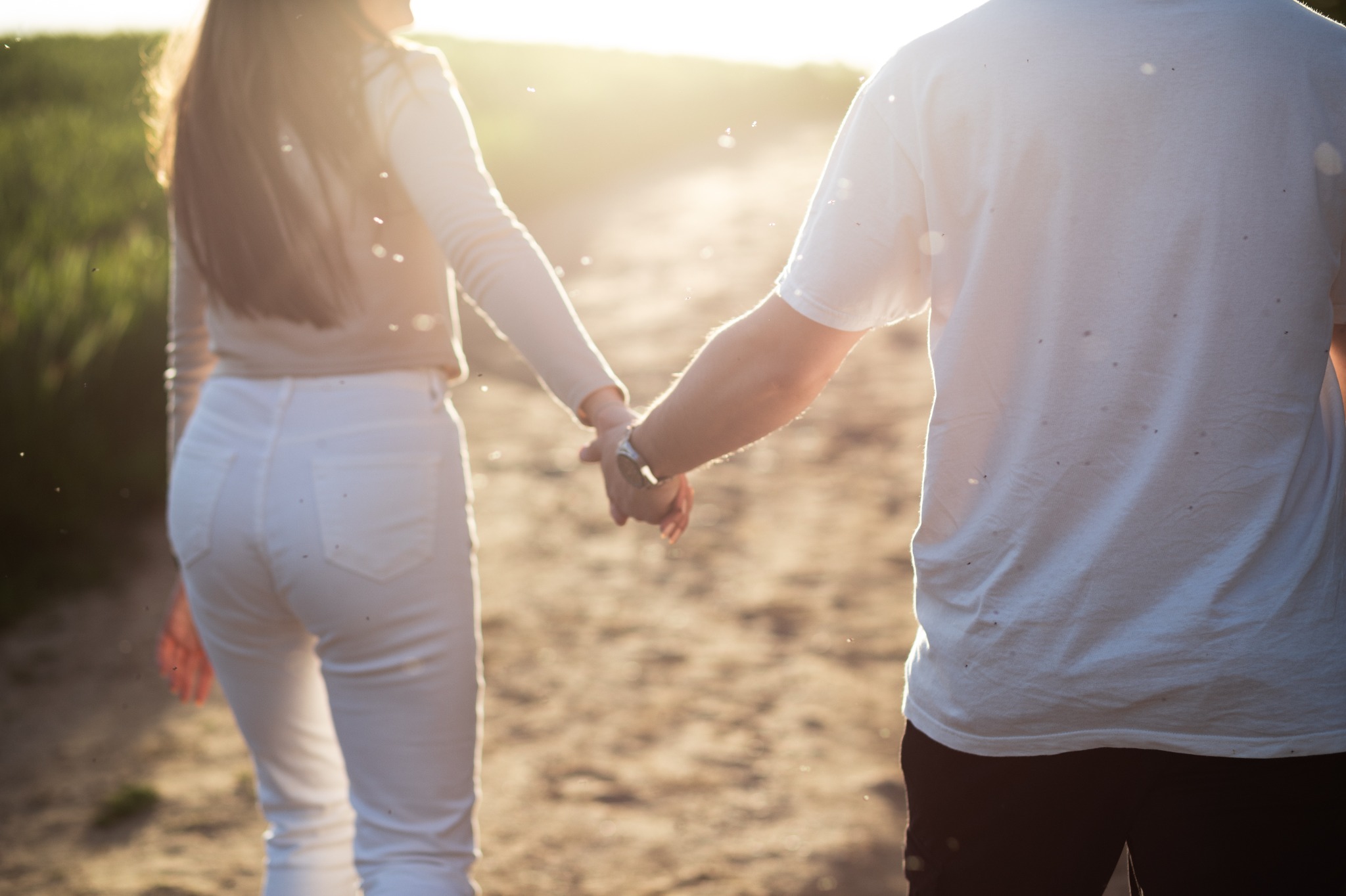 Para trzymająca się za ręce, idąca polną drogą w promieniach zachodzącego słońca. Kobieta w białych spodniach, mężczyzna w białym T-shircie. Widok z tyłu, z lekkim efektem bokeh.