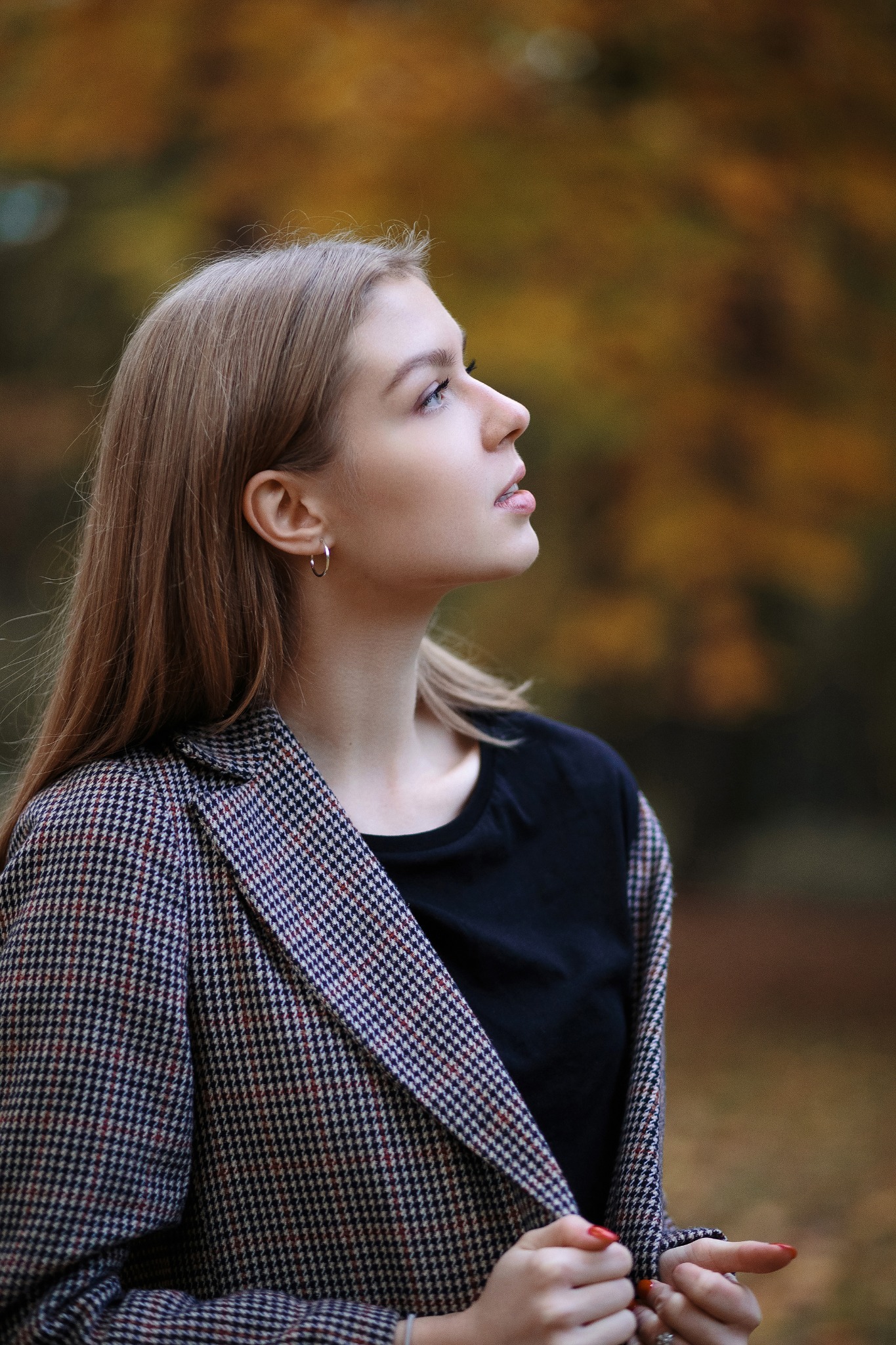 Portret młodej kobiety z profilu, w jesiennej scenerii, ubrana w marynarkę w pepitkę, z delikatnym makijażem i srebrnymi kolczykami-kołami, spoglądającej w górę.