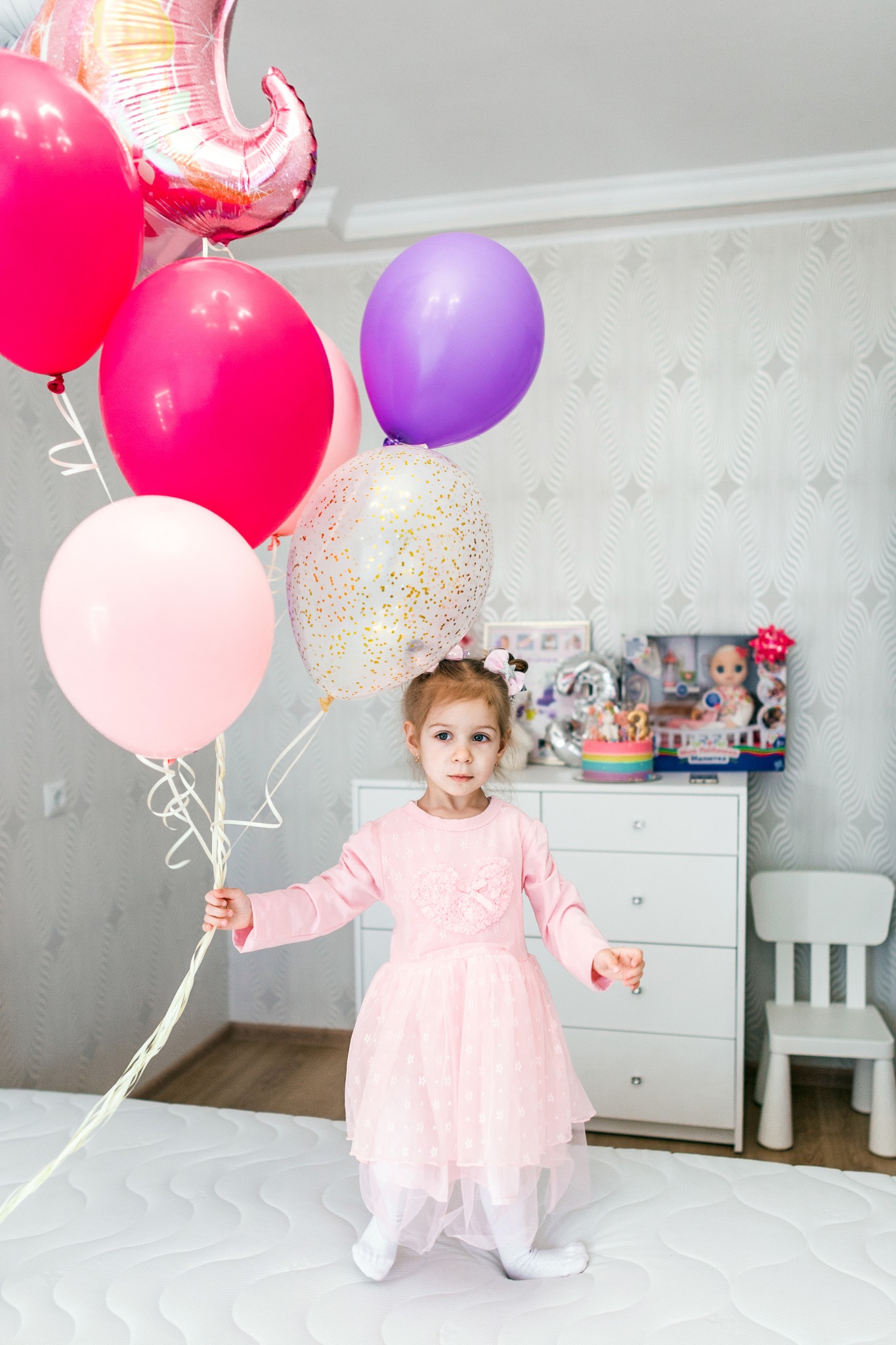 Uśmiechnięta dziewczynka w różowej sukience trzyma bukiet kolorowych balonów na tle pokoju z tortem urodzinowym i prezentami.