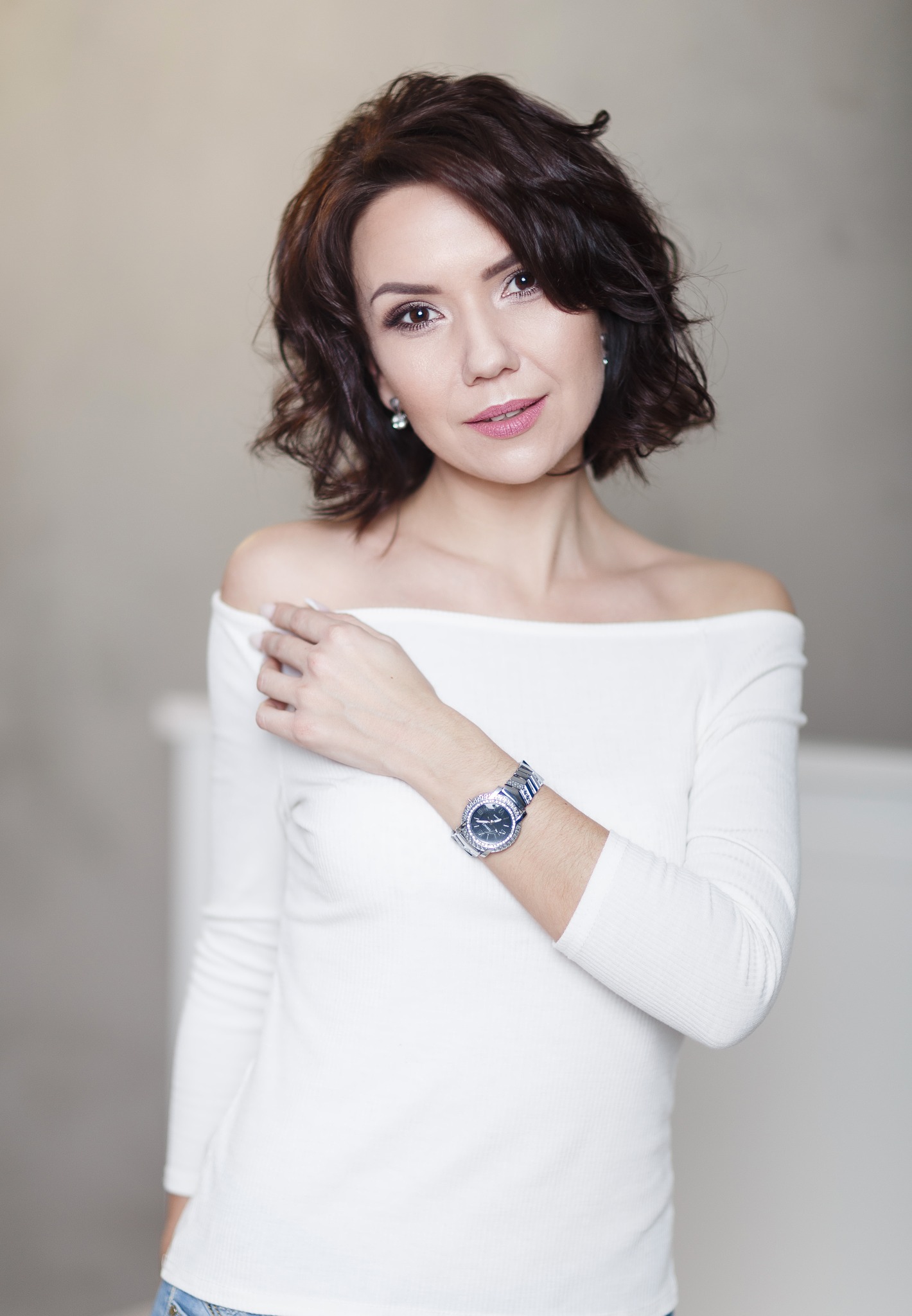 Portret kobiety z brązowymi, kręconymi włosami, ubranej w białą bluzkę z odkrytymi ramionami i srebrny zegarek na ręce, pozującej na rozmytym, jasnym tle.